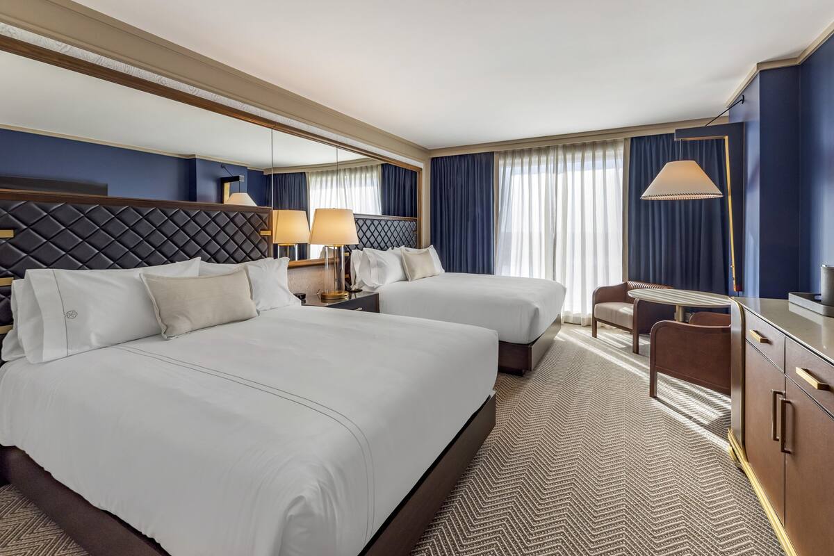 A hotel room with two beds, a desk, a chair, a lamp, and a large mirror.