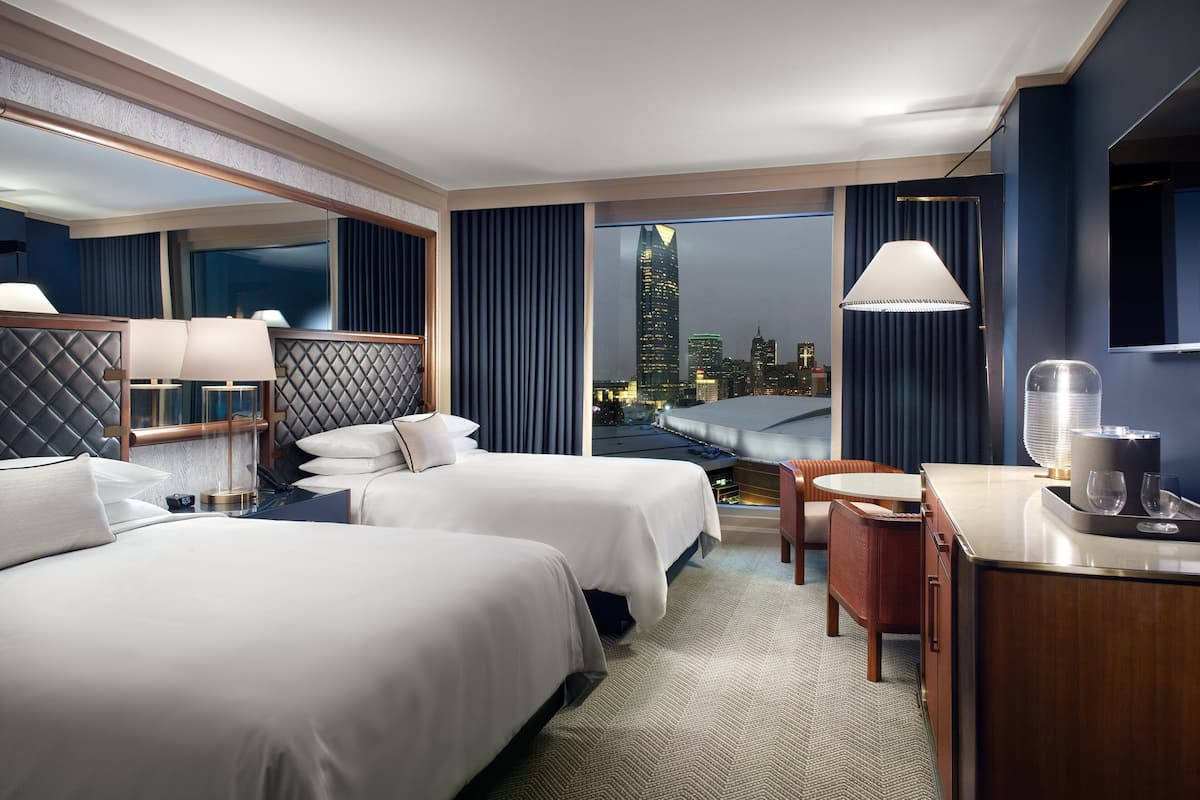 A hotel room with two beds, a desk with a chair, and a view of the city skyline at night.