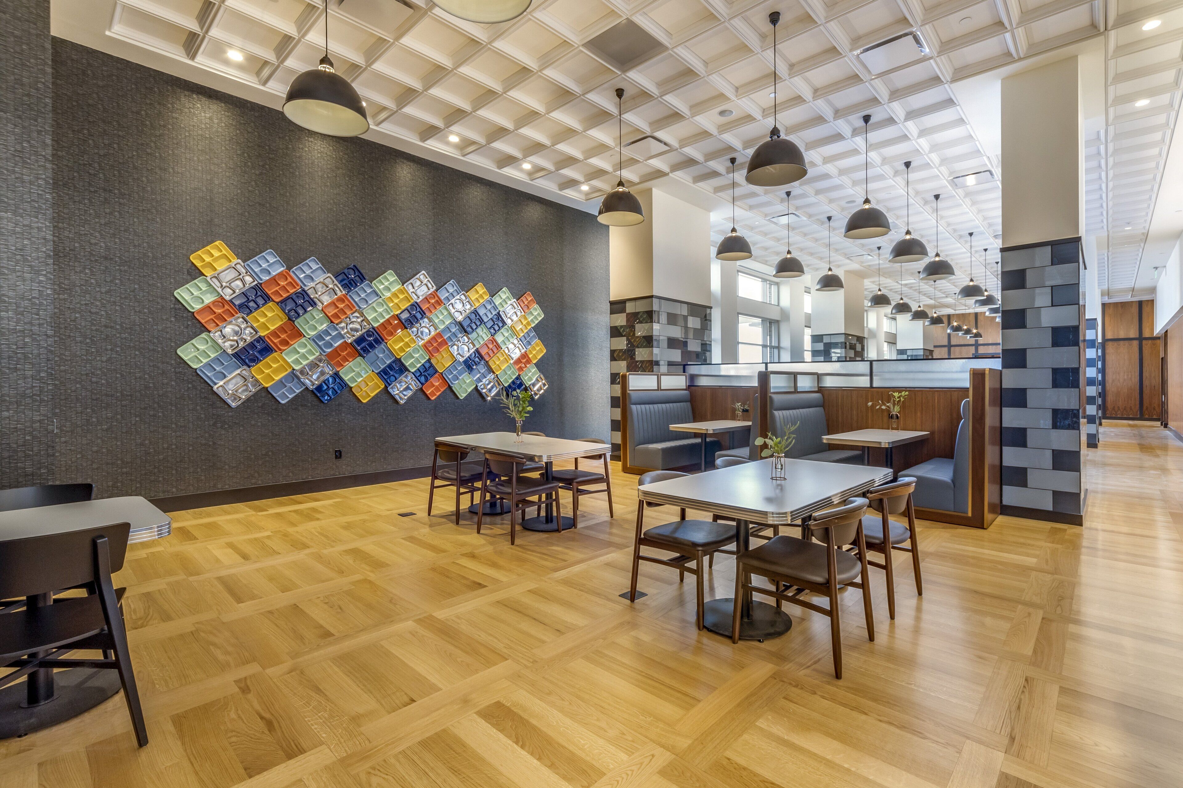 Ein modernes Restaurant mit Holzboden, mehreren Tischen und Sitzgruppen, Pendelleuchten und einer farbenfrohen Mosaikwand.