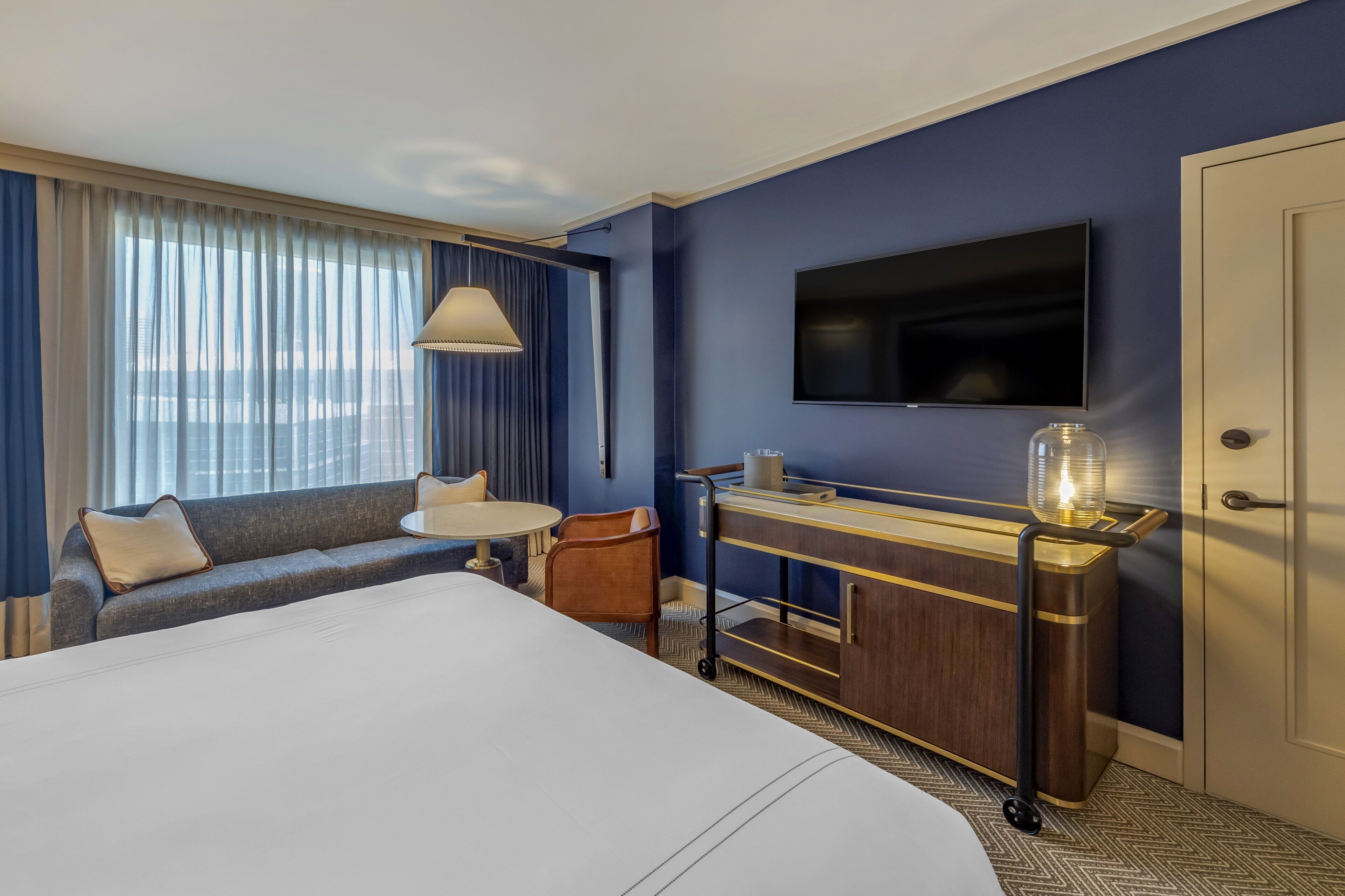 A hotel room with a large bed, a sofa, a small table, a TV, a minibar, and a view of the city through the window.