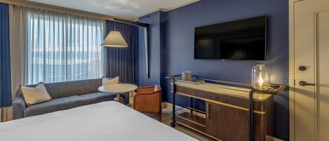 Habitación de hotel con una cama grande, un sofá, una mesita, televisión, minibar y vista a la ciudad desde la ventana.