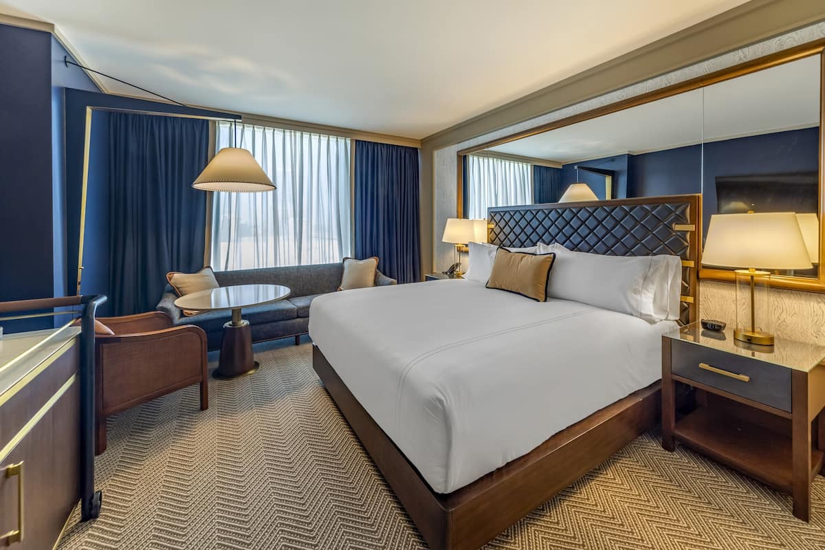A hotel room with a large bed, a sofa, a small round table, a desk, and a mirror.