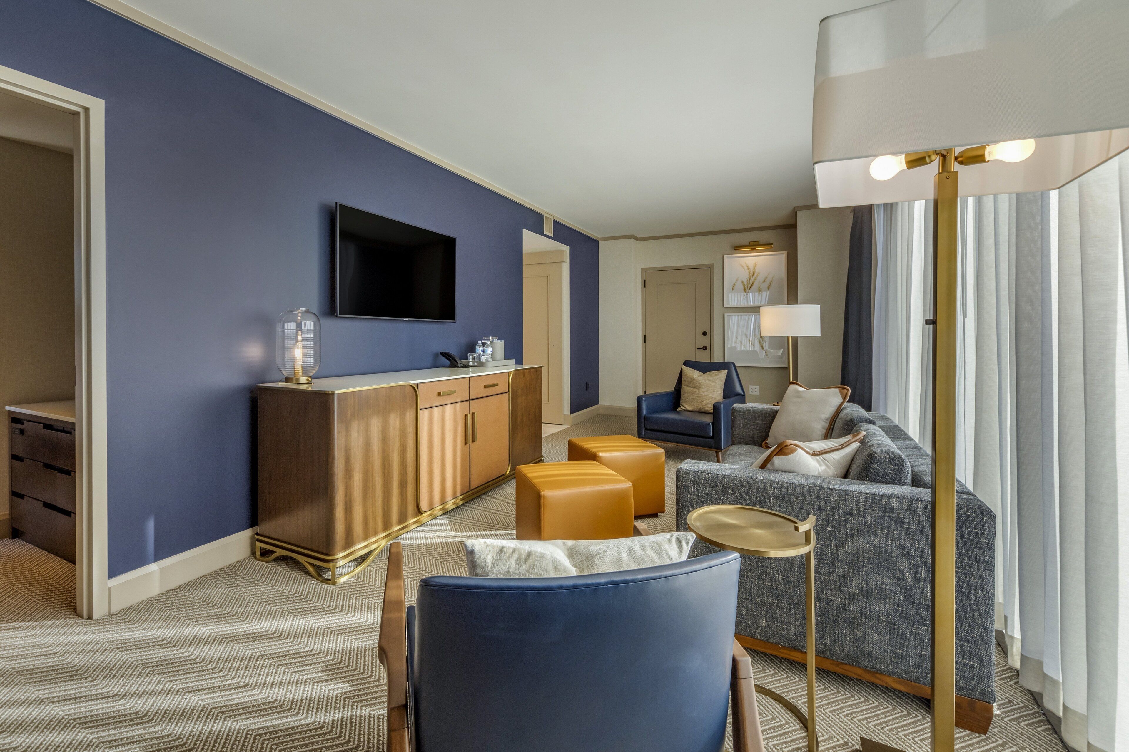 Una moderna camera d'albergo con una parete blu a contrasto, una TV a schermo piatto, un mobiletto in legno, un divano e una lampada da terra.