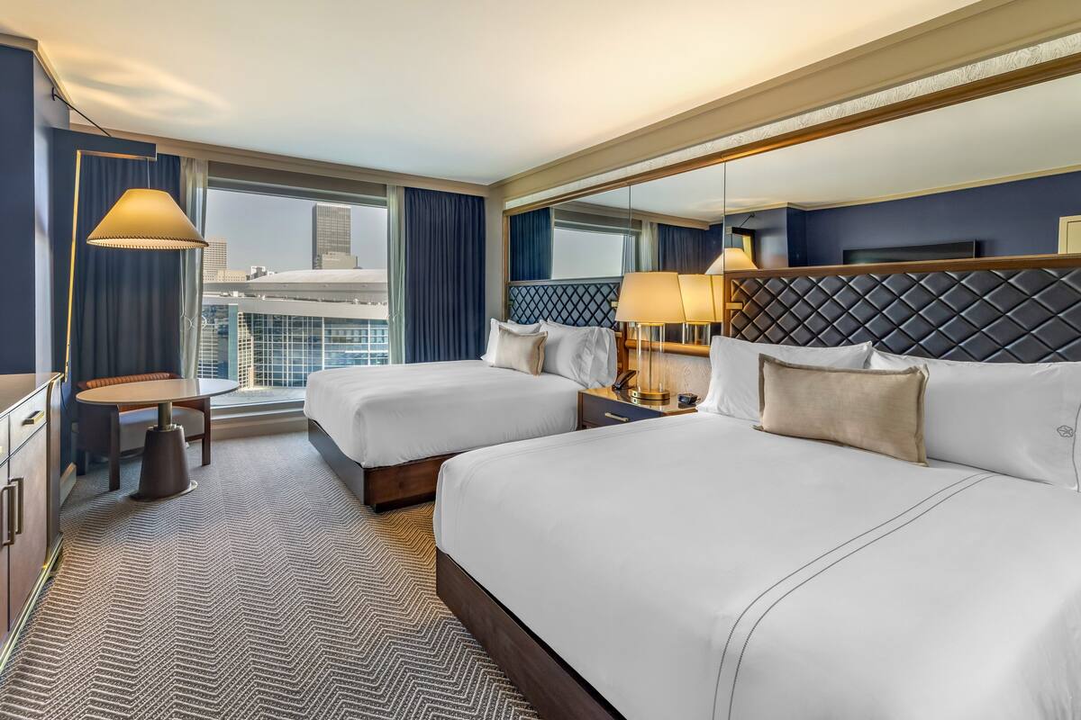 A hotel room with two beds, a large window with a city view, a small table, and a mirror above the bed.