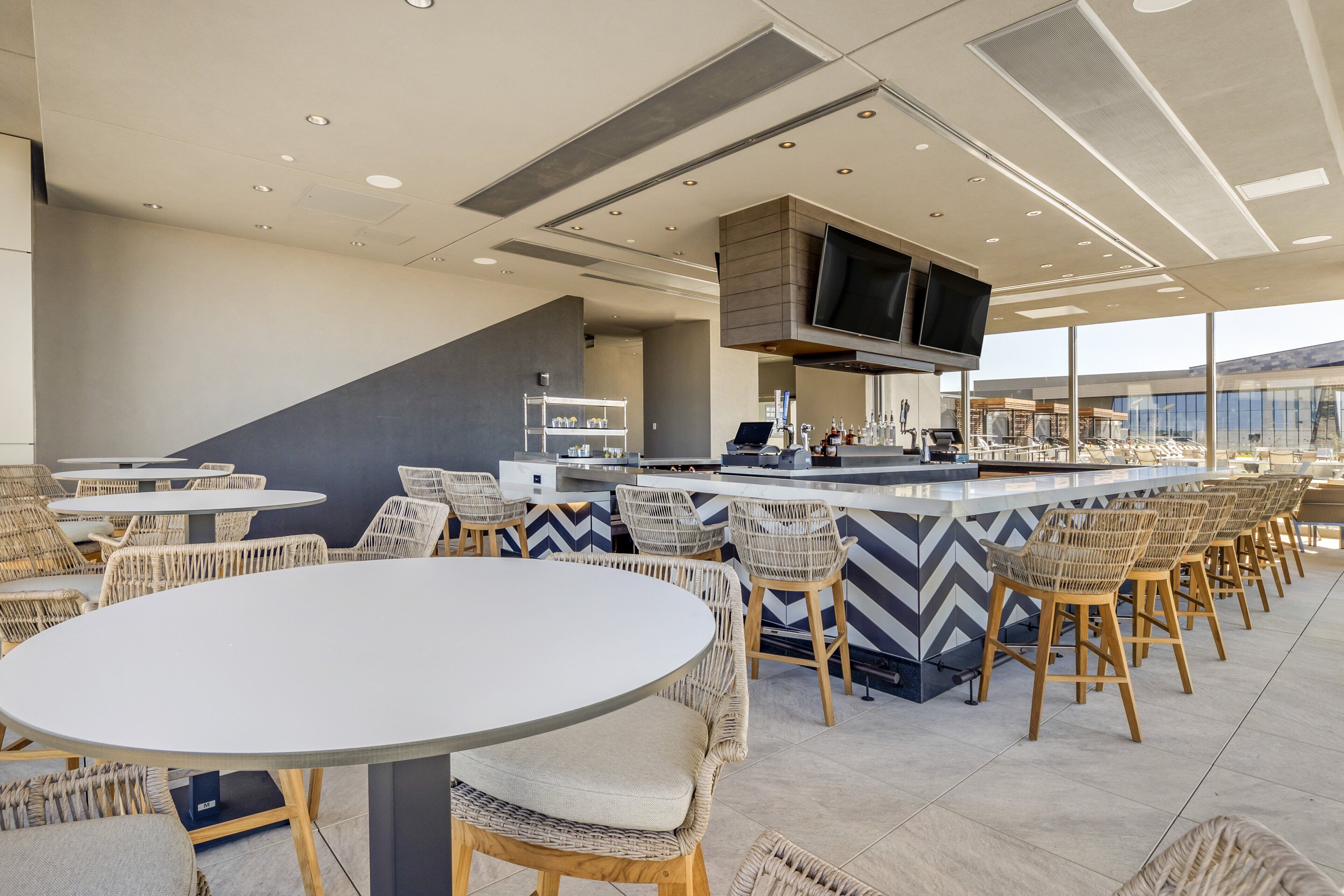A modern bar area with round tables and wicker chairs, a flat-screen TV mounted on the ceiling, and a bar counter with stools.