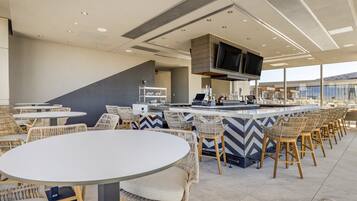 A modern bar area with round tables and wicker chairs, a flat-screen TV mounted on the ceiling, and a bar counter with stools.