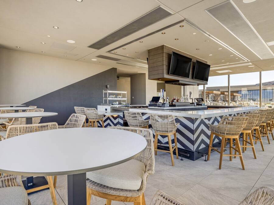 A modern bar area with round tables and wicker chairs, a flat-screen TV mounted on the ceiling, and a bar counter with stools.