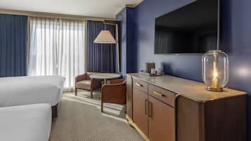 A hotel room with two beds, a desk with a lamp, a television, and a large window with curtains.