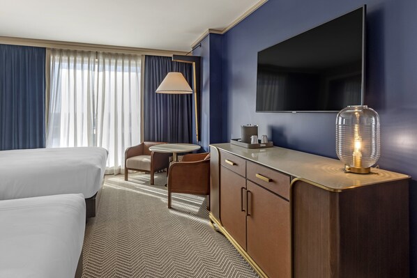 Una habitación de hotel con dos camas, un escritorio con lámpara, televisión y un ventanal con cortinas.