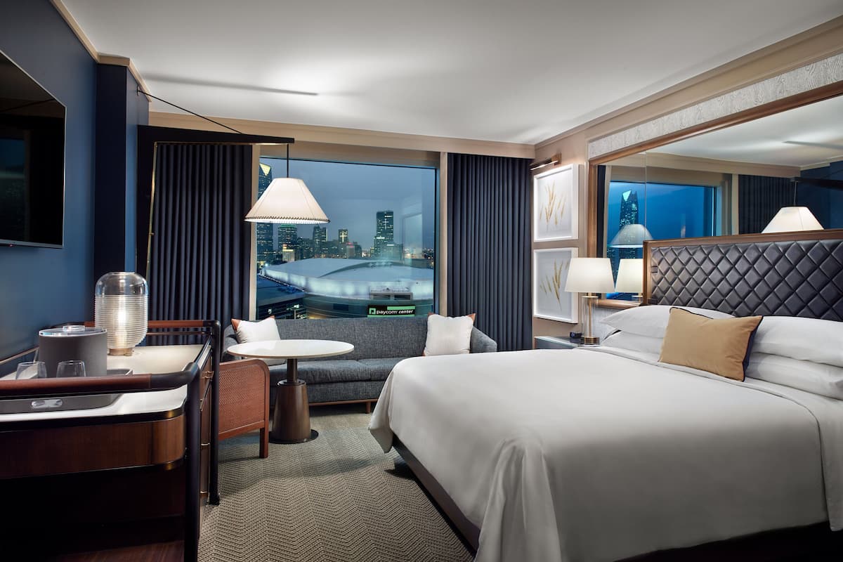 A hotel room with a large bed, a desk with a lamp, a chair, and a view of a cityscape.