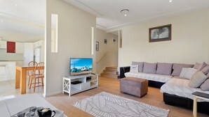 A modern living room with a TV, sofa, and coffee table. There is a staircase leading to another level. - Sea Change on Excellent (Vincentia)