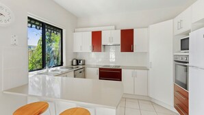 A modern kitchen with white cabinets, a central island, and a window with a view of greenery. - Sea Change on Excellent (Vincentia)