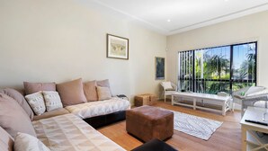 A modern living room with a sofa, ottoman, and a dining area with a table and chairs. There is a large window with a view of palm trees. - Sea Change on Excellent (Vincentia)