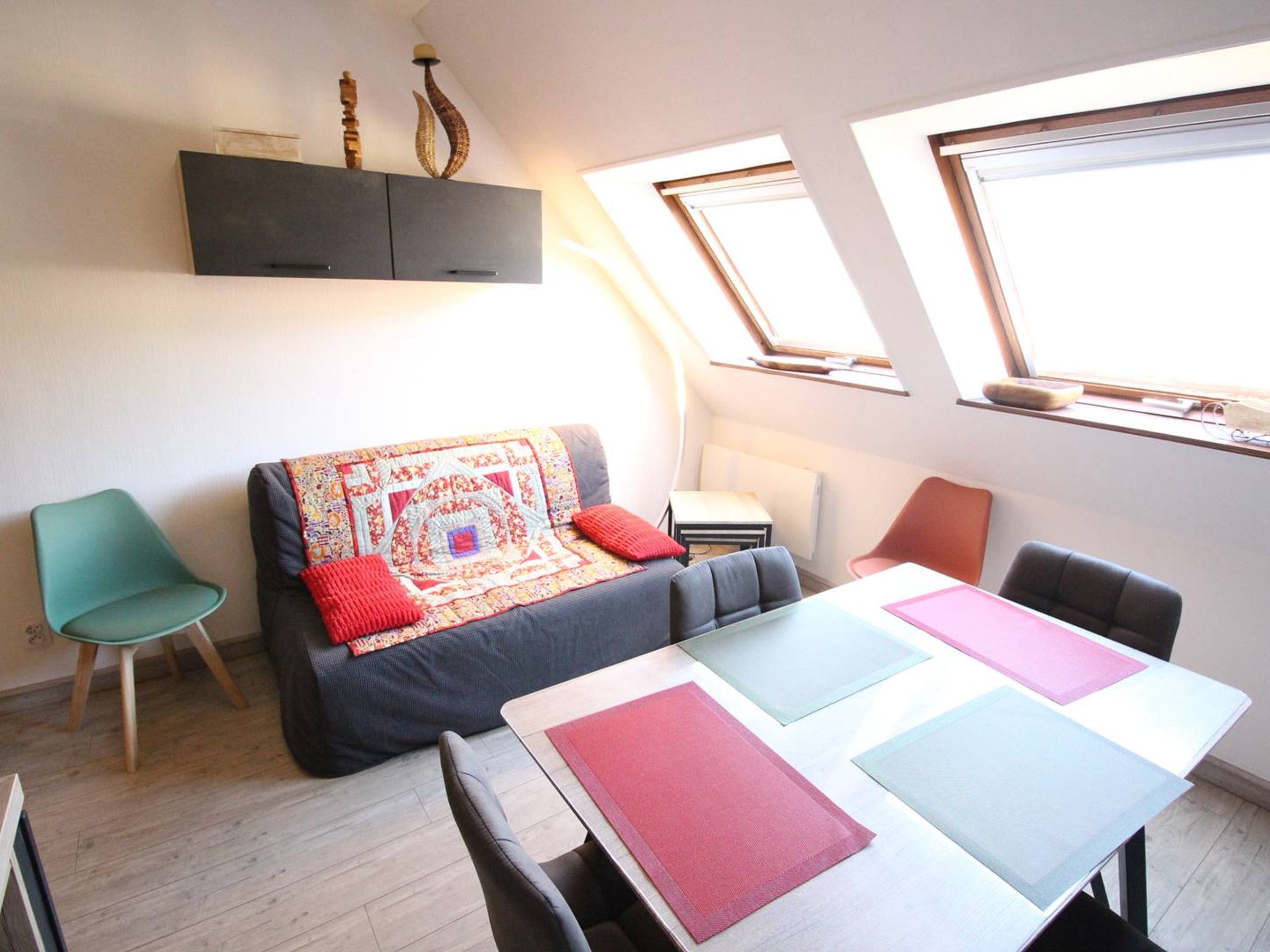 Ein kompakter Wohnraum mit einer Couch, einem Esstisch und einem kleinen Beistelltisch. Zwei Fenster sorgen für Tageslicht, und eine Decke mit einem Oberlicht.