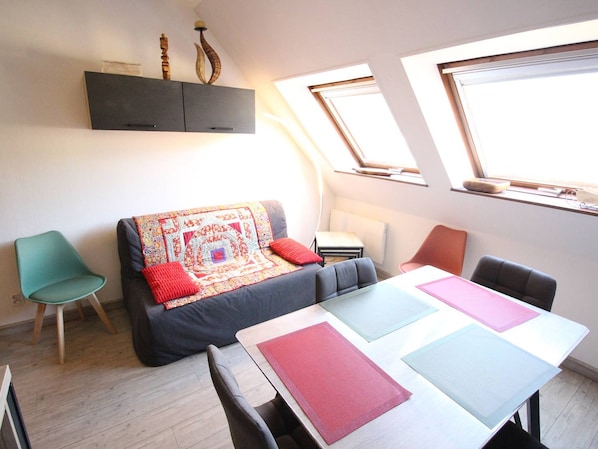 Ein kompakter Wohnraum mit einer Couch, einem Esstisch und einem kleinen Beistelltisch. Zwei Fenster sorgen für Tageslicht, und eine Decke mit einem Oberlicht.
