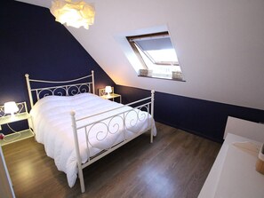 Ein Schlafzimmer mit schräg verlaufender Decke, einem Oberlicht, einem weißen Metallbettgestell, einem Nachttisch mit Lampe und Holzboden.