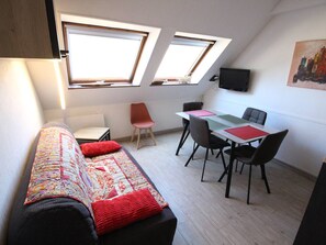 Ein kompakter Wohnraum mit einer Couch, einem Esstisch und zwei Stühlen. Oberlichter befinden sich über den Fenstern, und ein Flachbildfernseher ist an der Wand montiert.