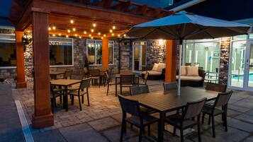An outdoor patio area with wooden tables and chairs, string lights, and a covered seating area with a large umbrella.
