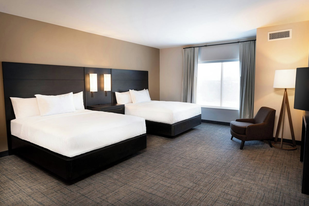 Habitación de hotel con dos camas, una silla, una lámpara y un ventanal grande.