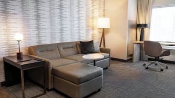 A modern hotel room with a sectional sofa, a small round table, a desk, a lamp, a chair, and a large window.