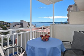 A balcony with a table covered in a blue tablecloth, a bowl of fruit, and a view of the sea and distant hills.