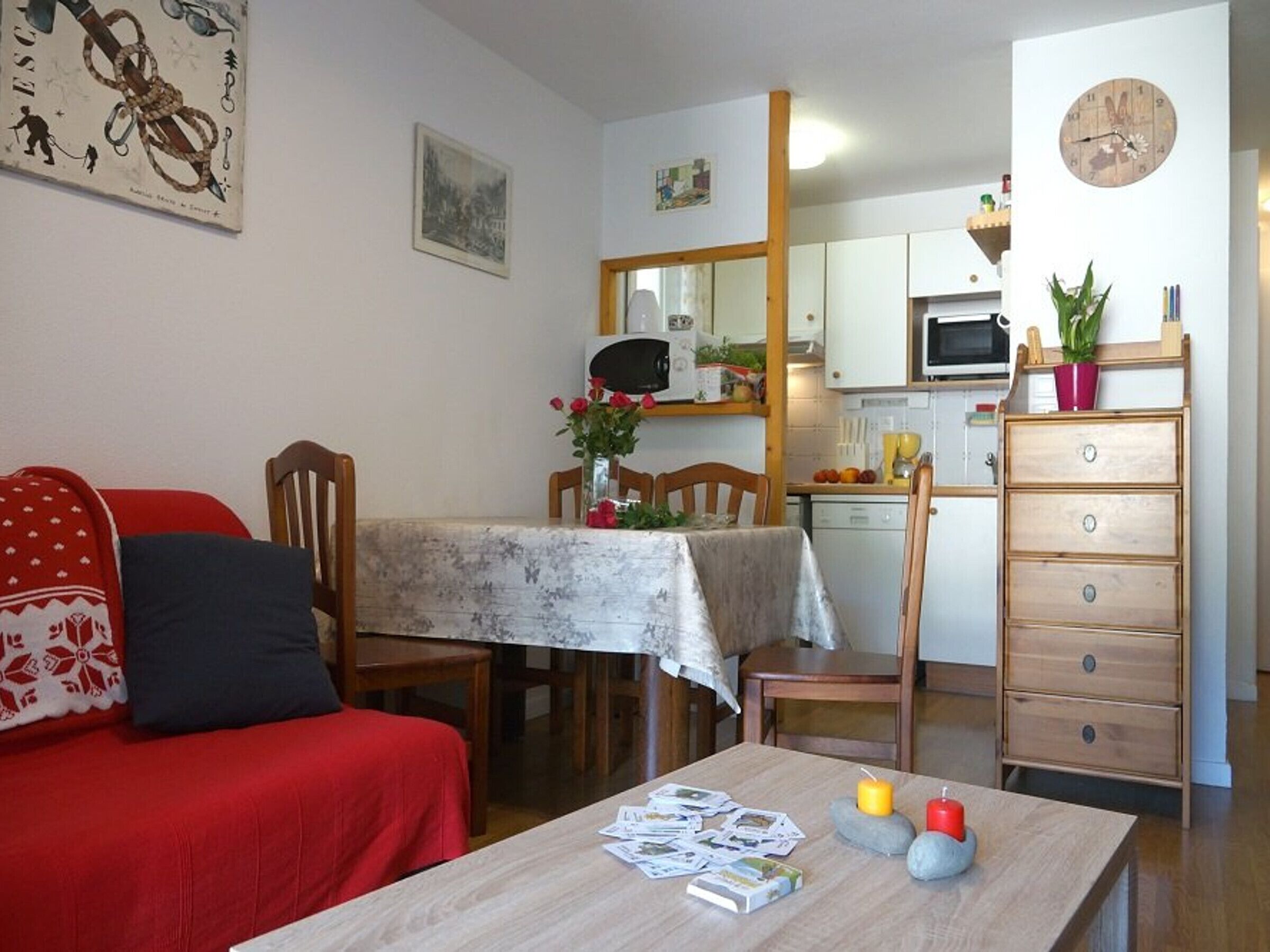 Una sala de estar acogedora con un sofá rojo, una mesa de comedor de madera con mantel floral, y una zona de cocina con armarios blancos y un microondas.