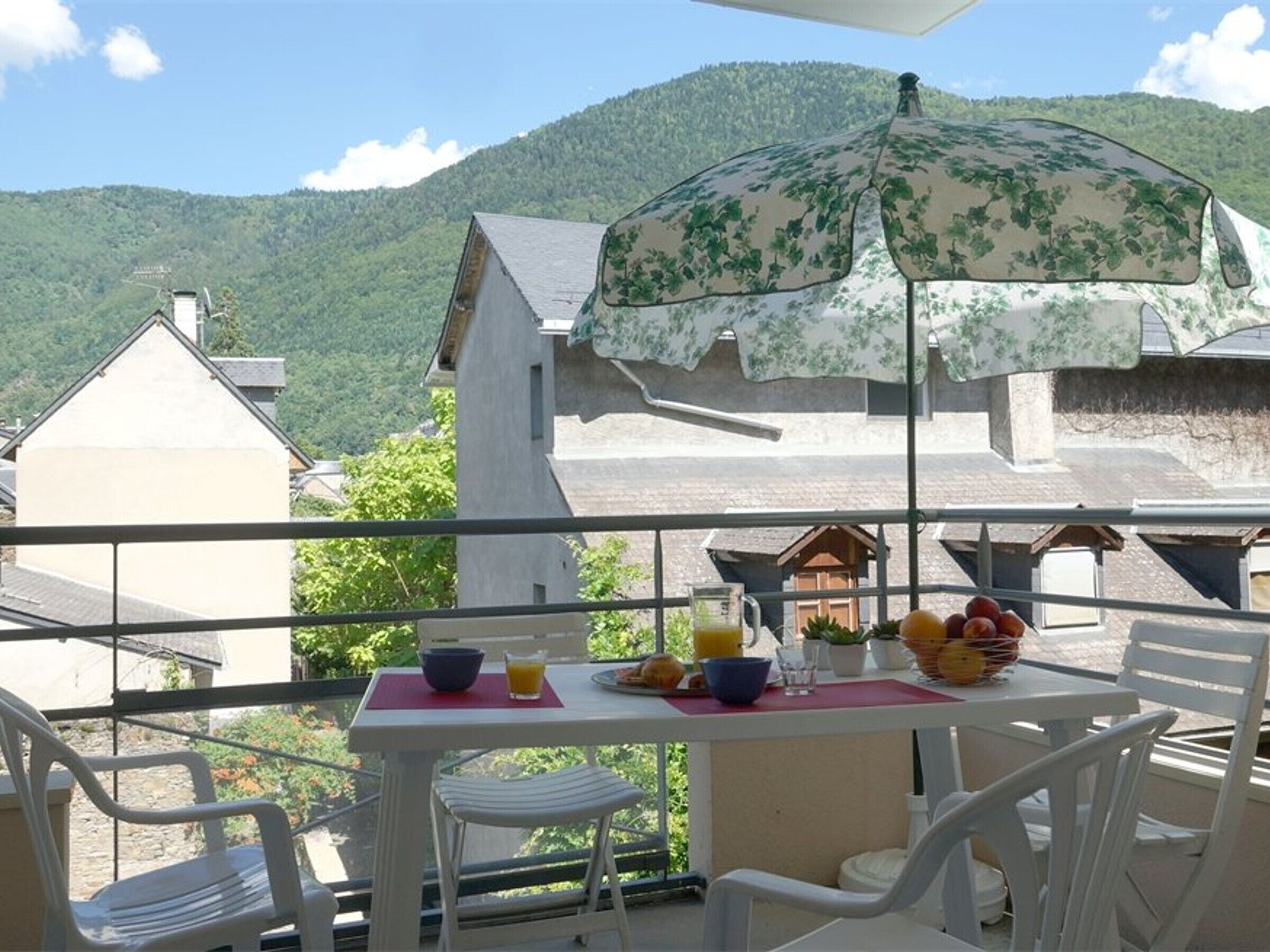 Un balcón con una mesa dispuesta para el desayuno, que incluye frutas y un vaso de zumo de naranja, con vistas a un paisaje montañoso.