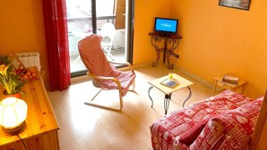 A cozy room with a rocking chair, a small table with a lamp, a bed with patterned bedding, a TV on a stand, and a window with curtains. - Central Luchon Studio for 2 - Balcony - Near Gondola (Bagnères-de-Luchon)