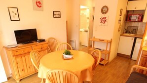 A cozy living room with a wooden dining table, chairs, a TV on a wooden cabinet, and a kitchen area with a microwave and refrigerator. - Near Thermal Baths - T2 with Balcony, Parking and WiFi - Pets Allowed (Bagnères-de-Luchon)