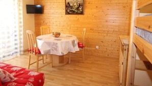 A cozy room with a wooden wall, a round table with chairs, a television mounted above the wall, and a bunk bed to the right. - Luchon: Central Apt - Garage, Balcony, 4 Guests, Near Lift & Thermal Baths (Bagnères-de-Luchon)