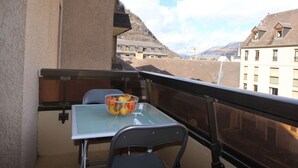 A balcony with a table and chairs, a fruit bowl, and a view of buildings and mountains. - Luchon: Central Apt - Garage, Balcony, 4 Guests, Near Lift & Thermal Baths (Bagnères-de-Luchon)