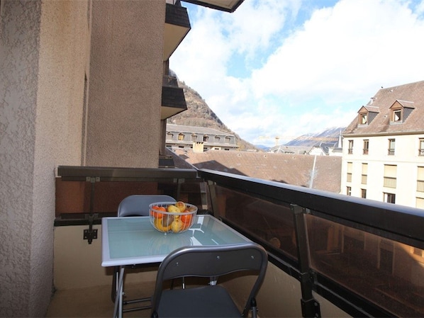 A balcony with a table and chairs, a fruit bowl, and a view of buildings and mountains.