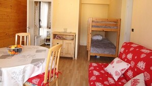 A compact room with a bunk bed, a small desk, a dining table with chairs, and a red sofa with patterned cushions. - Luchon: Central Apt - Garage, Balcony, 4 Guests, Near Lift & Thermal Baths (Bagnères-de-Luchon)