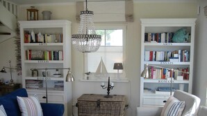 A cozy living room with a blue sofa, a checkered armchair, a wooden coffee table, and two white bookshelves labeled 'ARTS' and 'ROMANS'. A chandelier hangs from the ceiling, and a small table with a lamp is in the center. - Windrose, Deutschland (Breege)