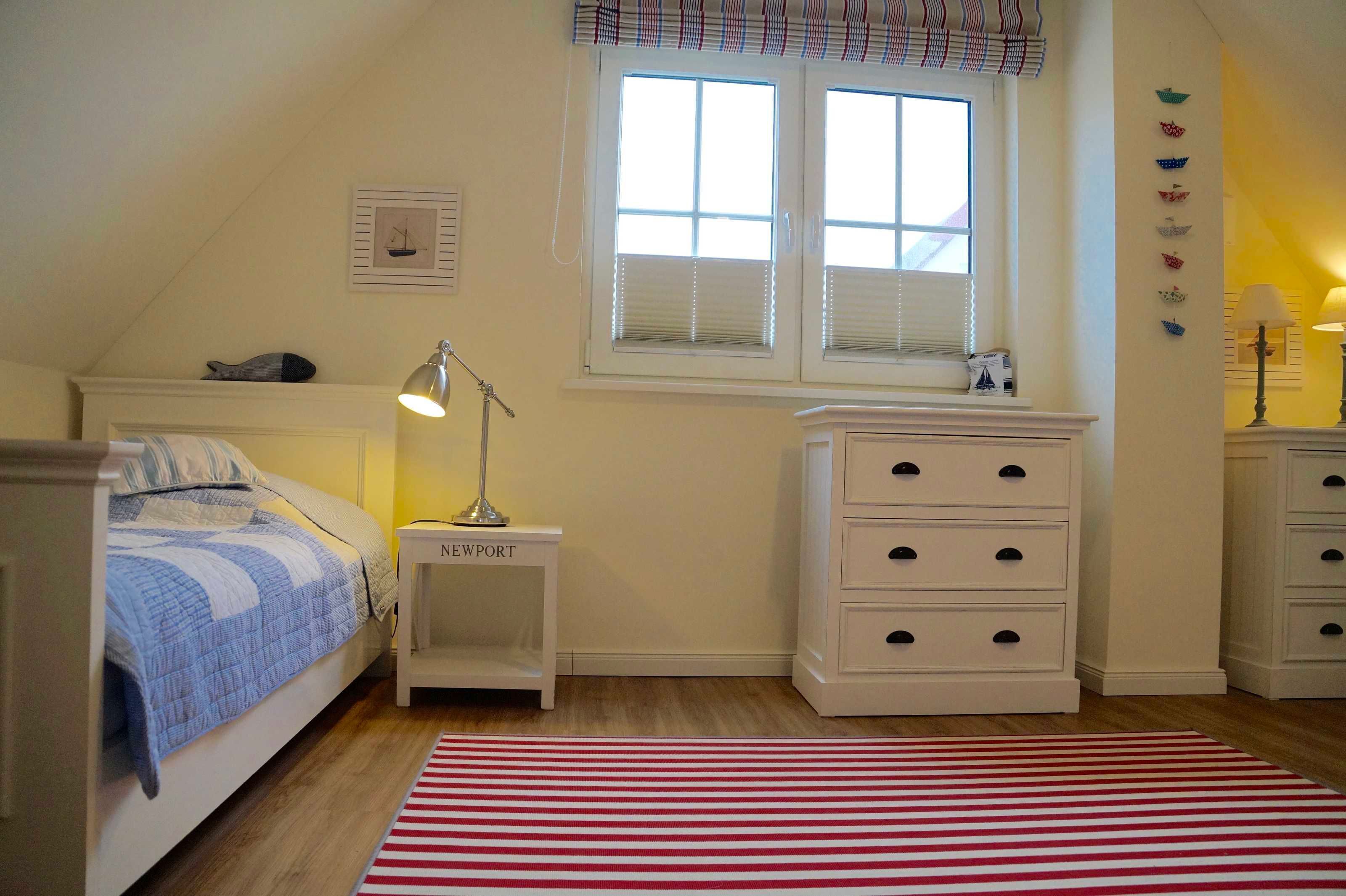 Quarto com cama, mesa de cabeceira, cômoda, janela com persianas e tapete listrado em vermelho e branco.
