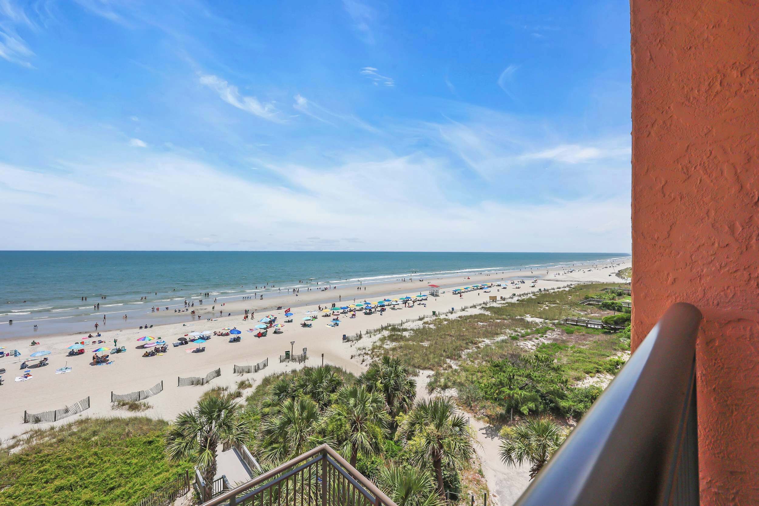 A beach view from a balcony with a railing, palm trees, and beach umbrellas.
