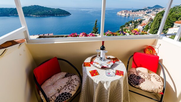 A balcony with a table set for two, featuring a bottle of wine, glasses, and a basket. The view includes a coastal town and a body of water. - Apartment Vedrana (Dubrovnik)