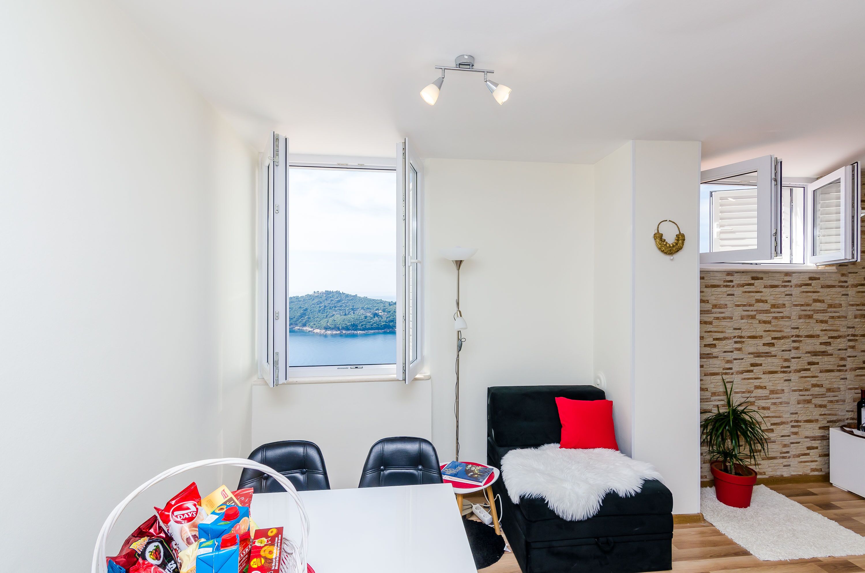A modern living room with a view of a hill and sea, a dining area with chairs and a table, and a black sofa with a red pillow.
