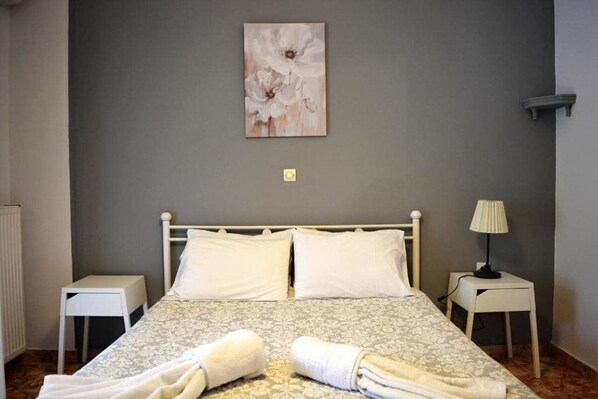 A neatly made bed with white linens and two rolled towels on the bed. There is a small bedside table with a lamp on the right side of the bed. The room has a gray wall with a framed artwork above the bed. - Despina Studio and Apartments (Corfu)