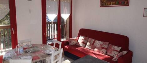 A cozy room with a red sofa, a dining table with chairs, and a large window with curtains.