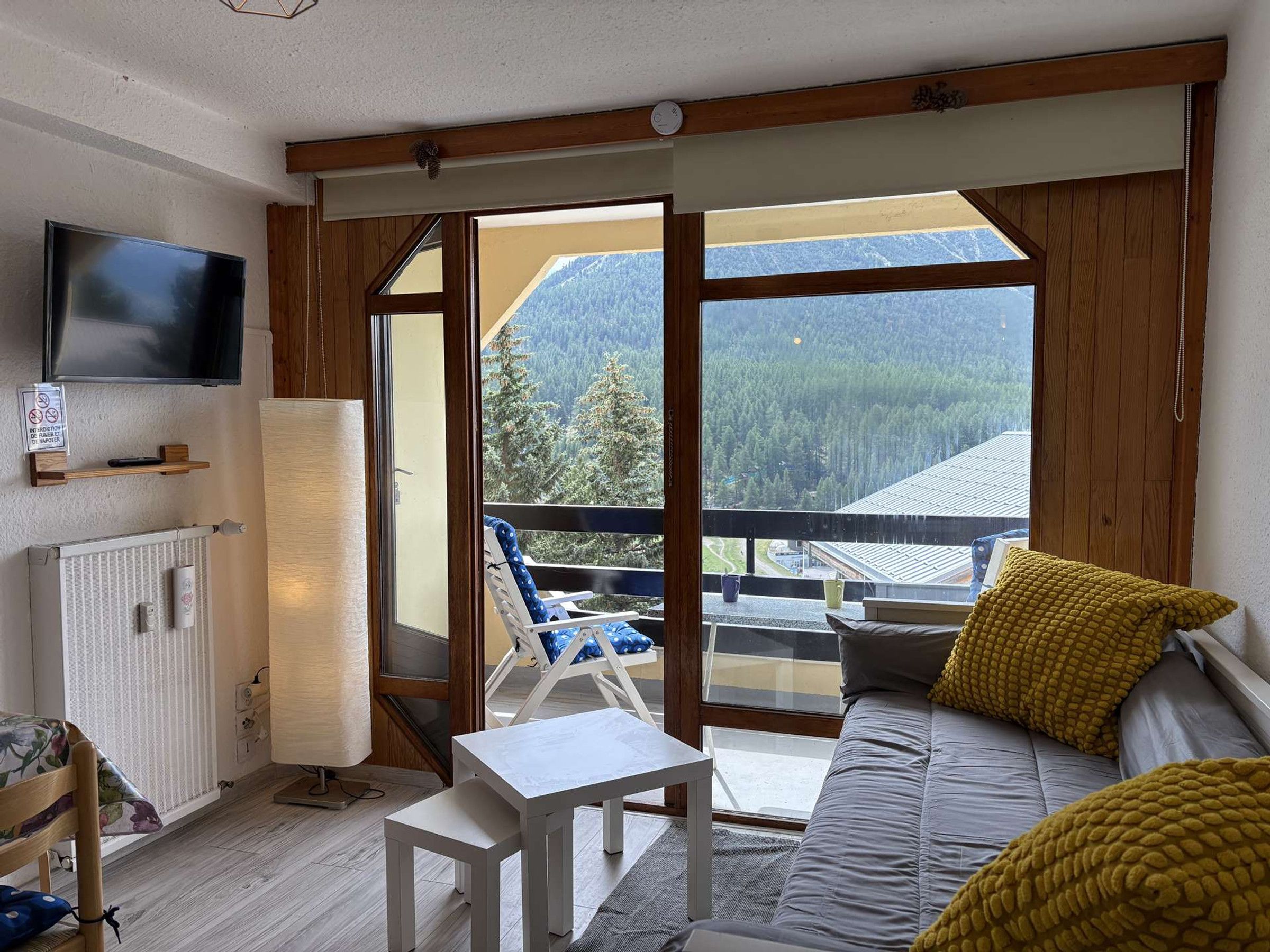 Une pièce comprenant un canapé, une petite table, un téléviseur fixé au mur et un balcon offrant une vue sur une montagne boisée.