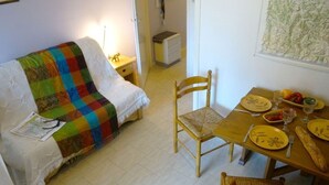 A cozy living room with a colorful quilted blanket on a white sofa, a wooden dining table set with yellow plates, and a wooden chair. - Peaceful South-Facing Studio in Luchon with Balcony, Parking, and Mountain View (Bagnères-de-Luchon)