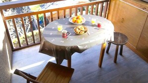 A round table with a floral tablecloth, a bowl of fruit, a mug, and a glass of orange juice on a balcony with wooden railings and a view of trees. - Peaceful South-Facing Studio in Luchon with Balcony, Parking, and Mountain View (Bagnères-de-Luchon)