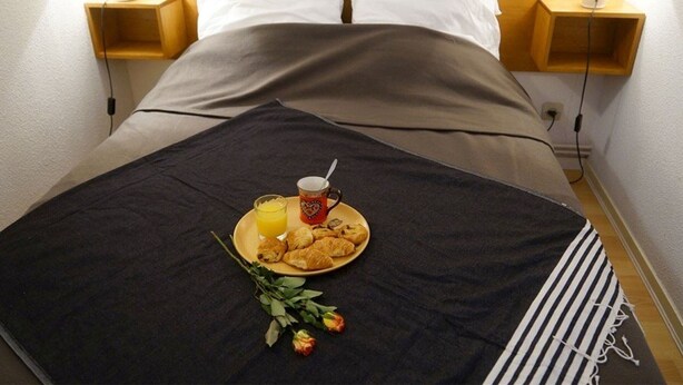 A neatly made bed with a dark blanket, a plate of croissants and orange juice, and a rose on the bed. - Peaceful South-Facing Studio in Luchon with Balcony, Parking, and Mountain View (Bagnères-de-Luchon)