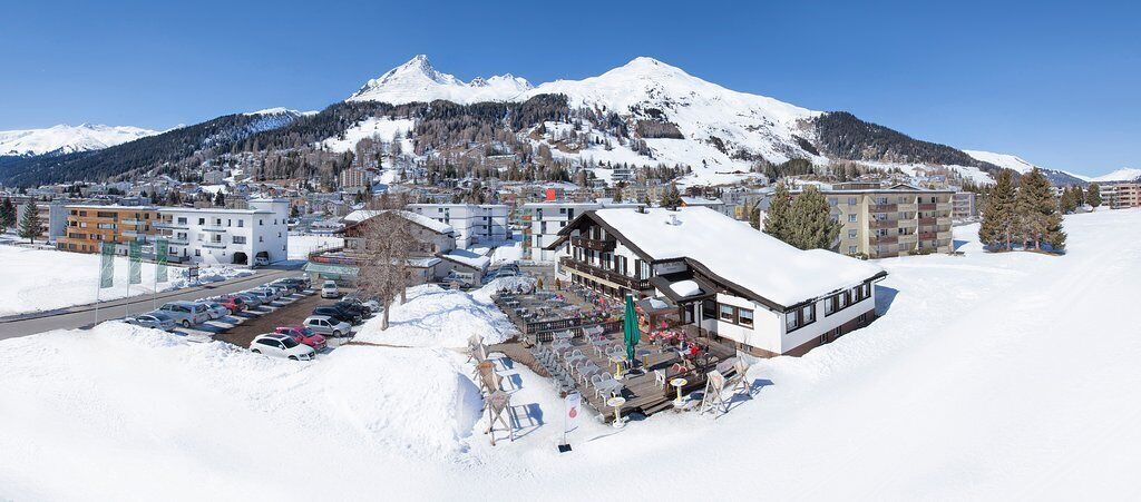 Un hotel con techo nevado, estacionamiento y montañas al fondo.
