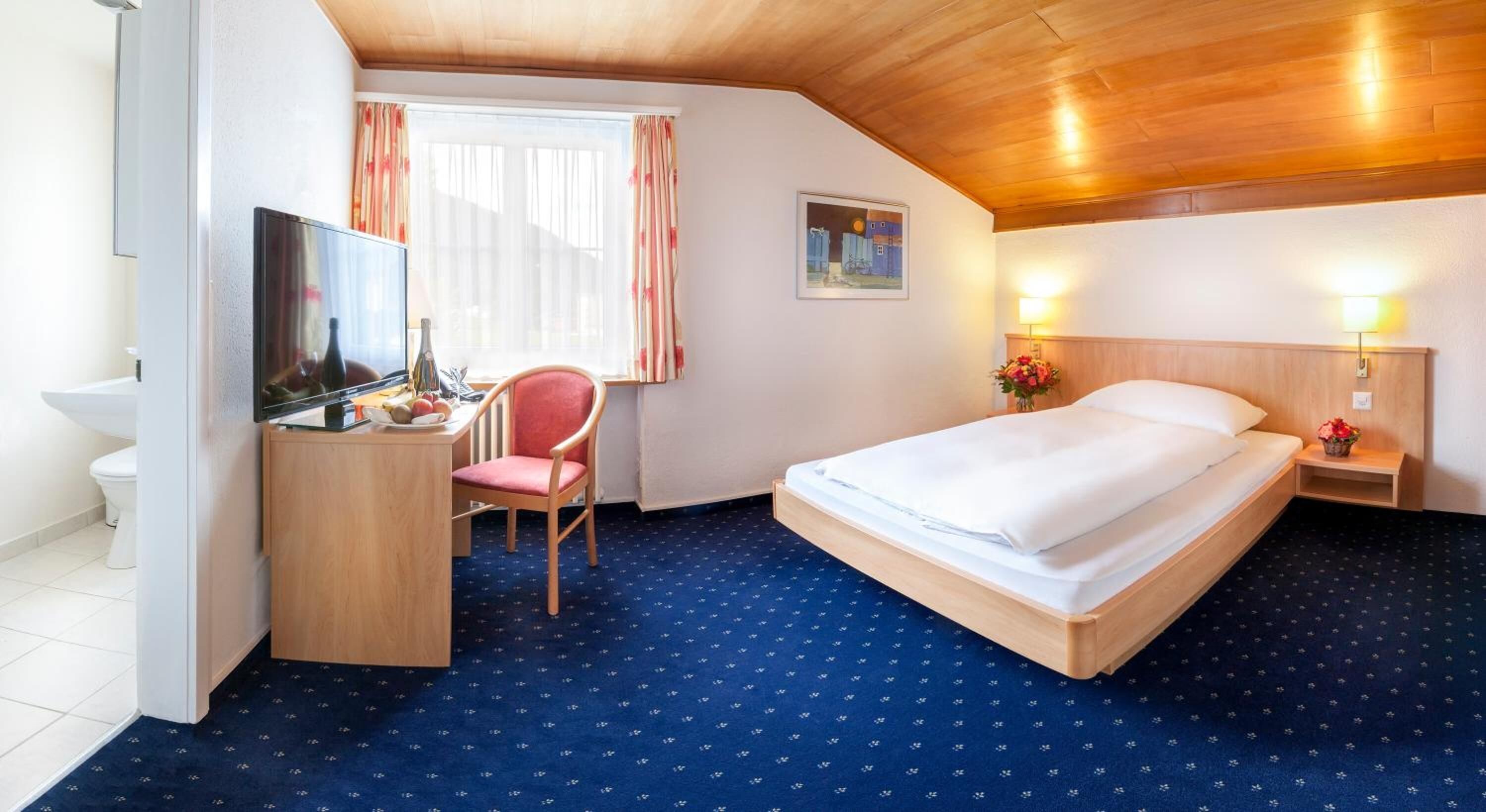 A hotel room with a bed, a desk with a TV, a chair, a window with curtains, and a painting on the wall.