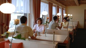 A restaurant with long communal tables, large windows with curtains, and hanging pendant lights. - Hotel Bünda Davos (Davos)