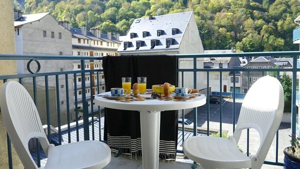 A balcony with two white chairs, a small round table with breakfast items, and a view of a forested hillside.