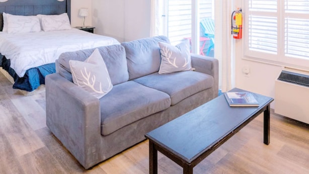 Une chambre à coucher avec un lit, un canapé gris, une petite table et une fenêtre avec des stores.