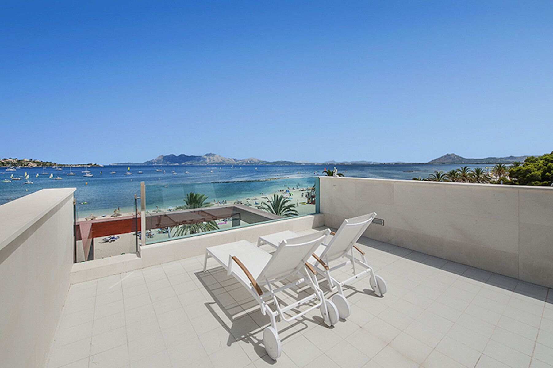 A terrace with white lounge chairs and a view of the sea and mountains.